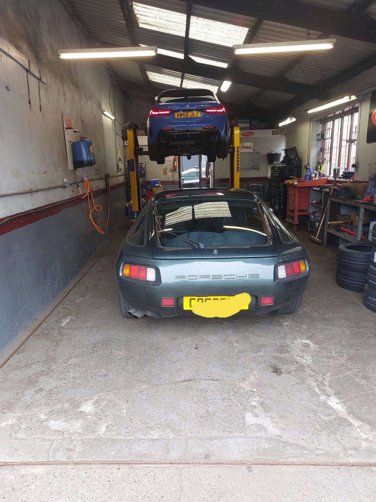 Porsche 928 in the bay with another car raised on the lift behind it